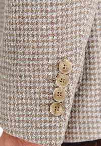 Brązowo-kremowy materiał w wzór houndstooth o teksturowanym wykończeniu, z czterema beżowymi guzikami na mankiecie rękawa.