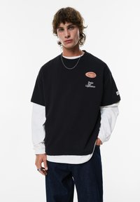 Ung mand med mellemlangt hår iført en lagdelt hvid langærmet skjorte og en sort kortærmet skjorte med tekst, mørke jeans og en sølv kæde halskæde.