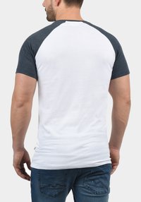 Bela majica s temno sivimi raglan rokavi, oprijeta zasnova, gladka bombažna tekstura in detajl stranskega šiva, kombinirana z modrimi kavbojkami.