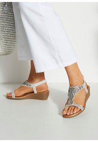 Sandalias de cuña en plata con detalles de cuentas, tiras planas y una suela texturizada color tostado. Combinadas con pantalones anchos blancos y un bolso de mimbre.