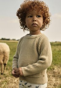 Beige, geripptes Strickoberteil mit langen Ärmeln, rundem Ausschnitt und seitlichen Schlitzen, getragen über gemusterten hellen Hosen. Weicher, strukturierter Stoff.