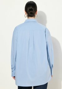 Camicia a maniche lunghe azzurro chiaro con colletto, dettaglio a pieghe sul retro e polsini con bottoni. Tessuto morbido con una vestibilità rilassata e oversize.