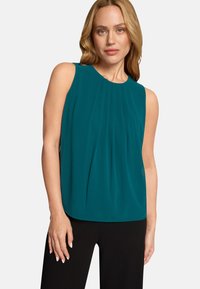Blouse sans manches couleur sarcelle avec détail plissé à l'avant, encolure ronde et tissu doux, associée à un pantalon noir. Texture lisse avec une coupe décontractée.