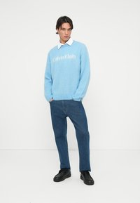 Fénykék, bolyhos pulóver, amelyen a "Calvin Klein" felirat található a mellrészen, sötétkék farmernadrággal és fekete cipővel párosítva. Alatta fehér galléros ing.