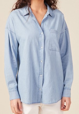 Kobieta w jasnoniebieskiej dżinsowej koszuli na guziki z długimi rękawami i kieszenią na piersi, w połączeniu z białymi spodniami.
