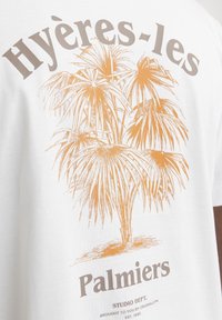 Witte T-shirt met een oranje palmbomenafbeelding en de tekst "Hyères-les Palmers" in bruin, geplaatst op de achterkant.