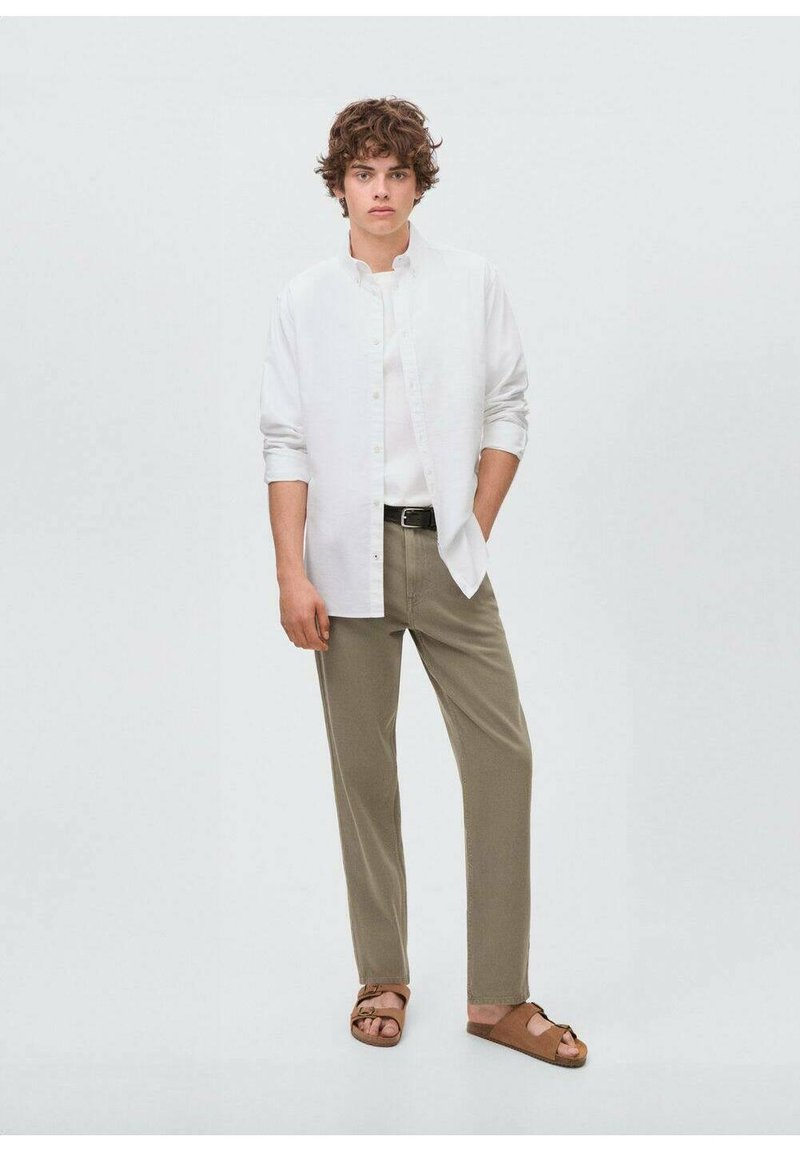 Camicia bianca a maniche lunghe, t-shirt bianca sporca, pantaloni verde oliva, sandali marroni. Il modello si trova in piedi con le mani in tasca contro uno sfondo neutro.