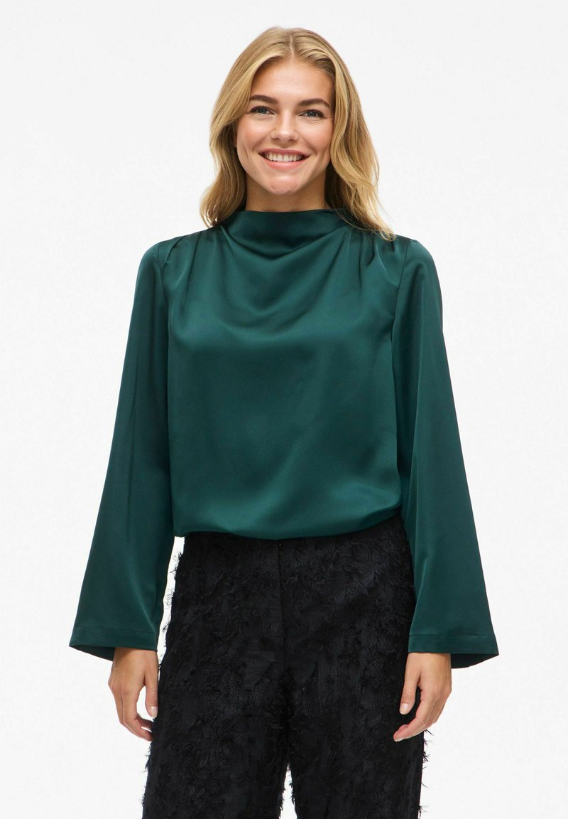 Grüne Satinbluse mit hohem Kragen und langen Ärmeln, die eine glatte Textur aufweist. Kombiniert mit schwarzen strukturierten Hosen.
