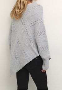 Kvinna som bär en ljusgrå stickad tröja med zigzag-mönster och spetsig nederkant, i kombination med svarta åtsittande byxor, sedd bakifrån.