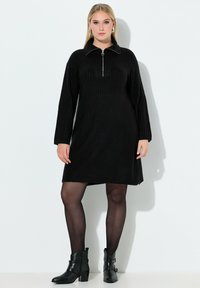 Zwarte geribbelde jurk met een ritssluiting bij de hals, lange mouwen en een losse pasvorm. Draagt zwarte enkellaarsjes en zwarte doorschijnende panty's.