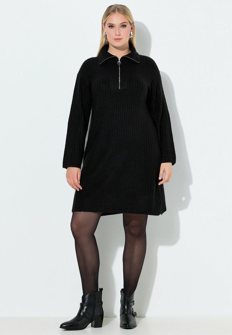 Zwarte geribbelde jurk met een ritssluiting bij de hals, lange mouwen en een losse pasvorm. Draagt zwarte enkellaarsjes en zwarte doorschijnende panty's.