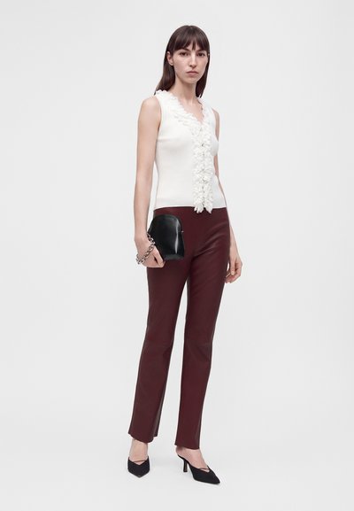 Femme portant un haut blanc sans manches avec des volants, un pantalon bordeaux, des chaussures noires à talons, et tenant un sac à main noir avec une bandoulière en chaîne.