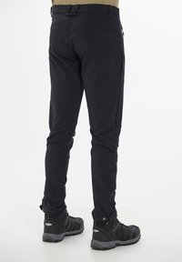 Svarta softshell-byxor med tapered fit, fickor med dragkedja på sidan och förstärkta muddar, i kombination med svarta utomhusskor.