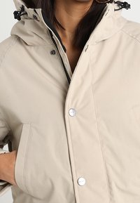 Chaqueta impermeable beige con capucha, que presenta cordones negros, botones a presión plateados y bolsillos laterales con una textura suave y mate.
