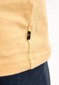 Bavlněné tričko ve světle žluté barvě s malou černou nášivkou se znakem na bočním švu; měkká textura s viditelným švem.