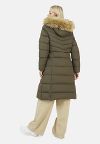 Cappotto imbottito verde oliva con cappuccio foderato in pelliccia, caratterizzato da cuciture a righe orizzontali e un design di lunghezza media. Indossato con pantaloni larghi beige.