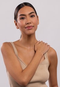 Zartes goldenes Armband mit kleinen runden Akzenten, an einem nackten Handgelenk getragen; das Model trägt ein braunes Rippenshirt und hat glatte Haut. Neutraler Hintergrund.