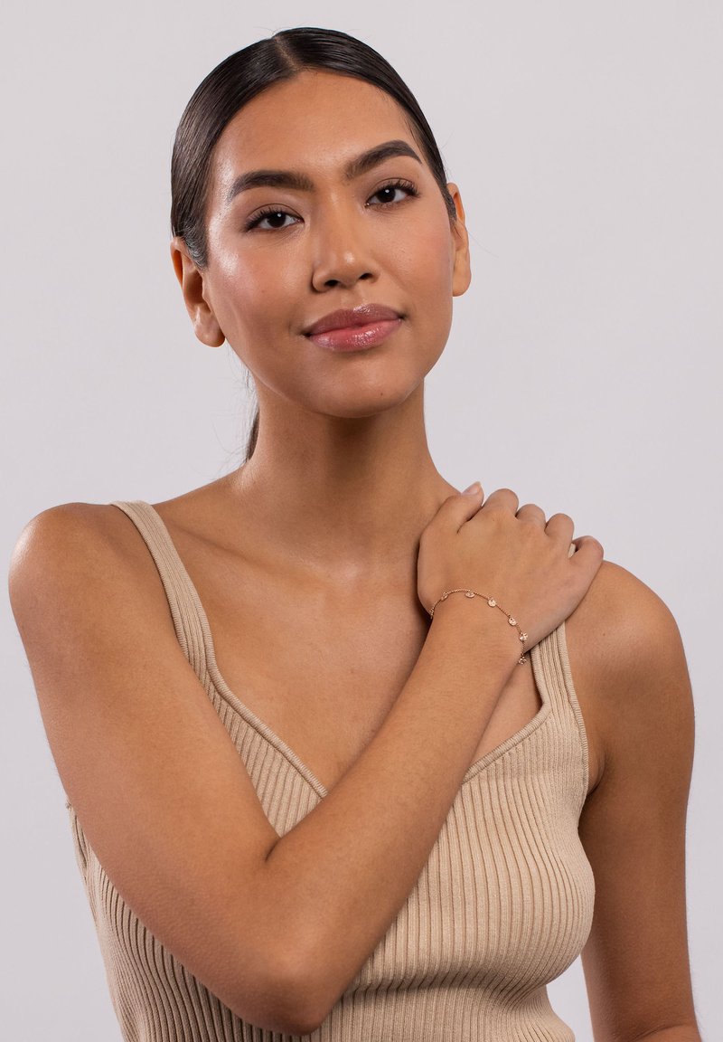 Zartes goldenes Armband mit kleinen runden Akzenten, an einem nackten Handgelenk getragen; das Model trägt ein braunes Rippenshirt und hat glatte Haut. Neutraler Hintergrund.