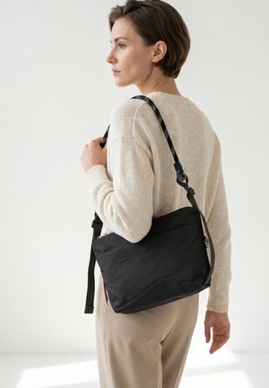 Donna che indossa un maglione color crema e pantaloni beige porta una borsa nera a tracolla con una tracolla regolabile sulla spalla.