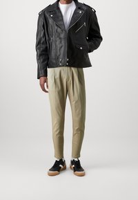 Muž v černé kožené moto bundě, bílém tričku, béžových skládacích kalhotách a černých teniskách s bílou a hnědou podrážkou, stojící na bílém pozadí.