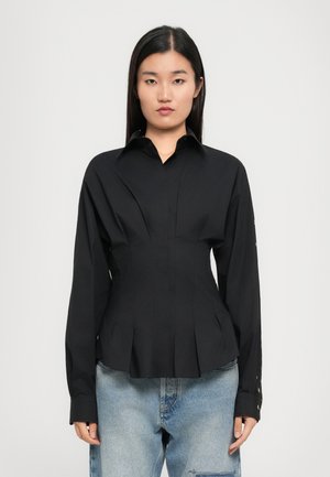 Vrouw met steil zwart haar draagt een strakke zwarte longsleeve shirt met plooien en lichtblauwe spijkerbroek, staande tegen een effen achtergrond.