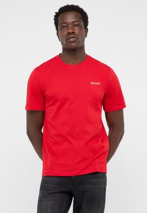 Mann steht mit den Händen hinter dem Rücken und trägt ein rotes kurzärmeliges T-Shirt mit kleinem weißem "Mustang" Text auf der linken Brust.