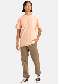 Camiseta de manga corta color melocotón claro con cuello redondo y un pequeño logo, combinada con pantalones cargo beige y zapatillas negras con suelas blancas.