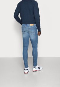 Vaqueros ajustados de color azul con un acabado desgastado, cinco bolsillos y una etiqueta marrón en la cintura trasera, combinados con zapatillas blancas.