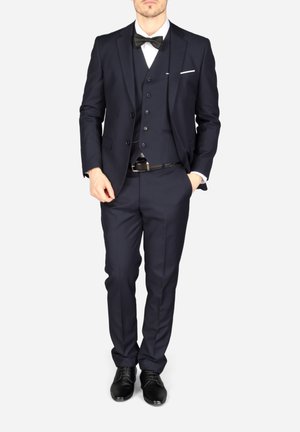 Man draagt een donker marineblauw driedelig pak met zwarte vlinderdas, wit overhemd, zwarte riem en zwarte nette schoenen, staand met handen gedeeltelijk in de zakken.