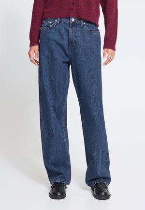 Personne portant un jean en denim bleu foncé ample, des chaussures noires et un haut à manches longues bordeaux, debout devant un fond clair uni.