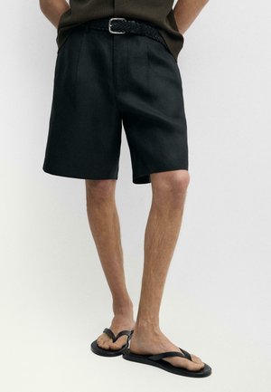 Mand iført sorte knælange shorts med et flettet sort bælte og sorte flip-flops, stående mod en ensfarvet hvid baggrund.