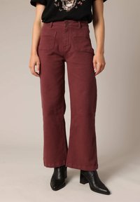 Pantalons larges bordeaux à taille haute, dotés de poches avant, d'une texture lisse et d'une fermeture par bouton, associés à des bottines noires.