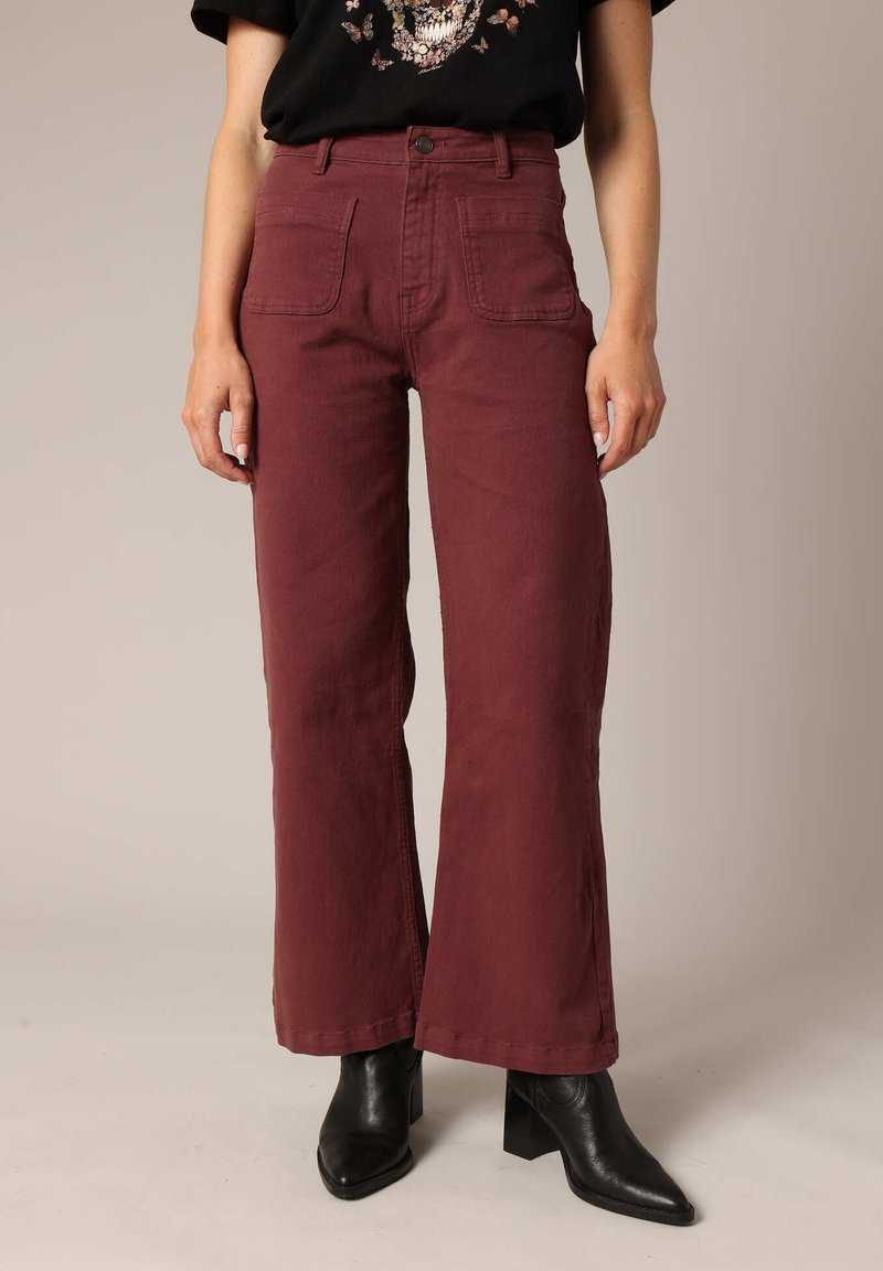 Pantalons larges bordeaux à taille haute, dotés de poches avant, d'une texture lisse et d'une fermeture par bouton, associés à des bottines noires.