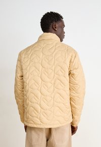 Beige vävd jacka med böjda sömmar och hög krage, med en avslappnad passform och elastiska ärmslut, bärs över ljusa byxor.