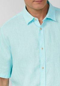 Camicia azzurra a maniche corte in lino, con colletto button-down e bottoni in legno lungo la patta. Texture liscia e design informale.