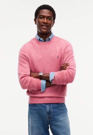 Man met gekruiste armen die een roze trui draagt over een blauw denim overhemd en blauwe spijkerbroek, glimlachend tegen een effen witte achtergrond.