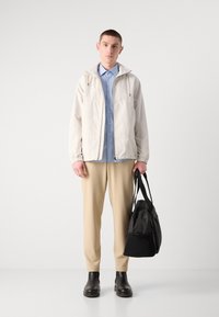 Lys beige jakke med en teksturert finish, lagdelt over en lyseblå skjorte. Kremfargede bukser og svarte støvler, holder en svart veske.