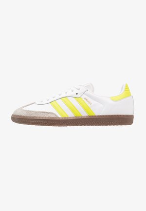 Baskets blanches Adidas Samba avec des bandes jaunes, une semelle gomme et un embout en daim beige, vues de côté.