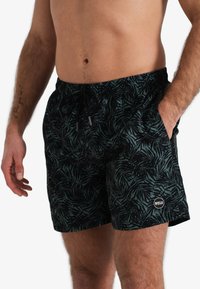 Zwarte zwemshorts met een donkergroene tropische bladerenpatroon, elastische tailleband en trekkoord. Voorzien van zijzakken en een logo-detail.