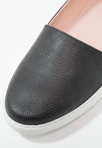 Chaussure noire sans lacets avec surface texturée et semelle en caoutchouc blanche, gros plan sur les détails avant et latéraux.