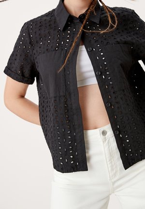 Femme portant une chemise noire à manches courtes ajourée boutonnée, par-dessus un débardeur blanc court et un pantalon blanc taille haute, vue des épaules aux hanches.