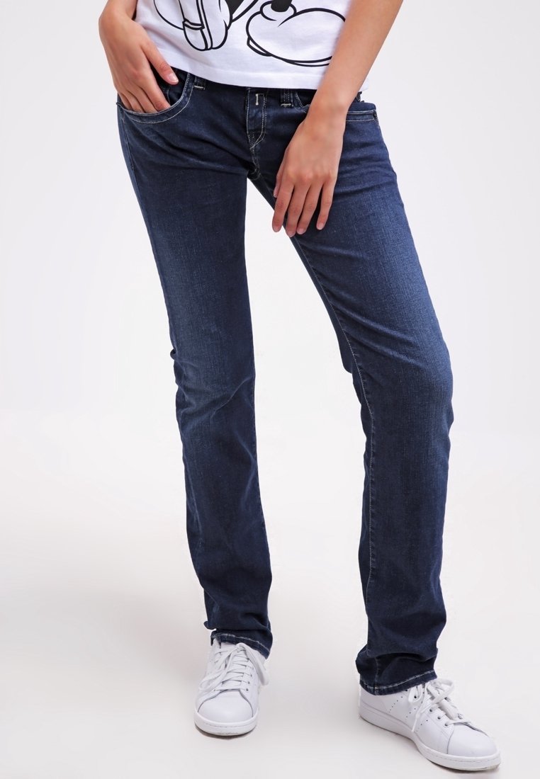 Jeans de denim azul oscuro con corte slim, diseño de cinco bolsillos, un ligero efecto de desgaste y un leve desgaste cerca de las rodillas. Zapatillas blancas puestas.