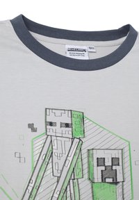 Camiseta de color gris claro con un cuello en contraste de color gris oscuro, que presenta un gráfico impreso de personajes pixelados en verde y negro.