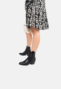 Botas de cuero negro hasta el tobillo con punta afilada y tacón cuadrado, que presentan una cremallera lateral y una tira para tirar. Combinadas con un vestido de estampado floral.