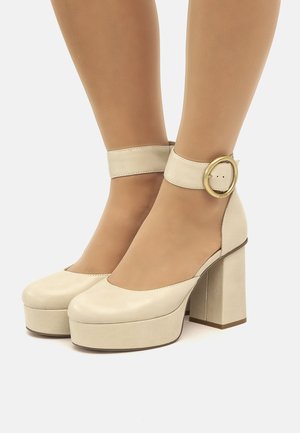 Tacones de plataforma beige con punta redondeada, correa para el tobillo y hebilla circular de oro. Fabricados en cuero suave con un tacón cuadrado grueso.