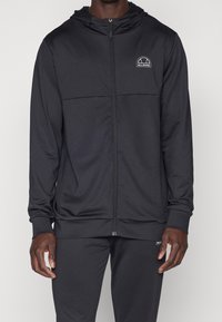 Haut à capuche zippé noir et pantalon assorti porté par une personne, avec le logo Ellesse sur la poitrine et la cuisse.