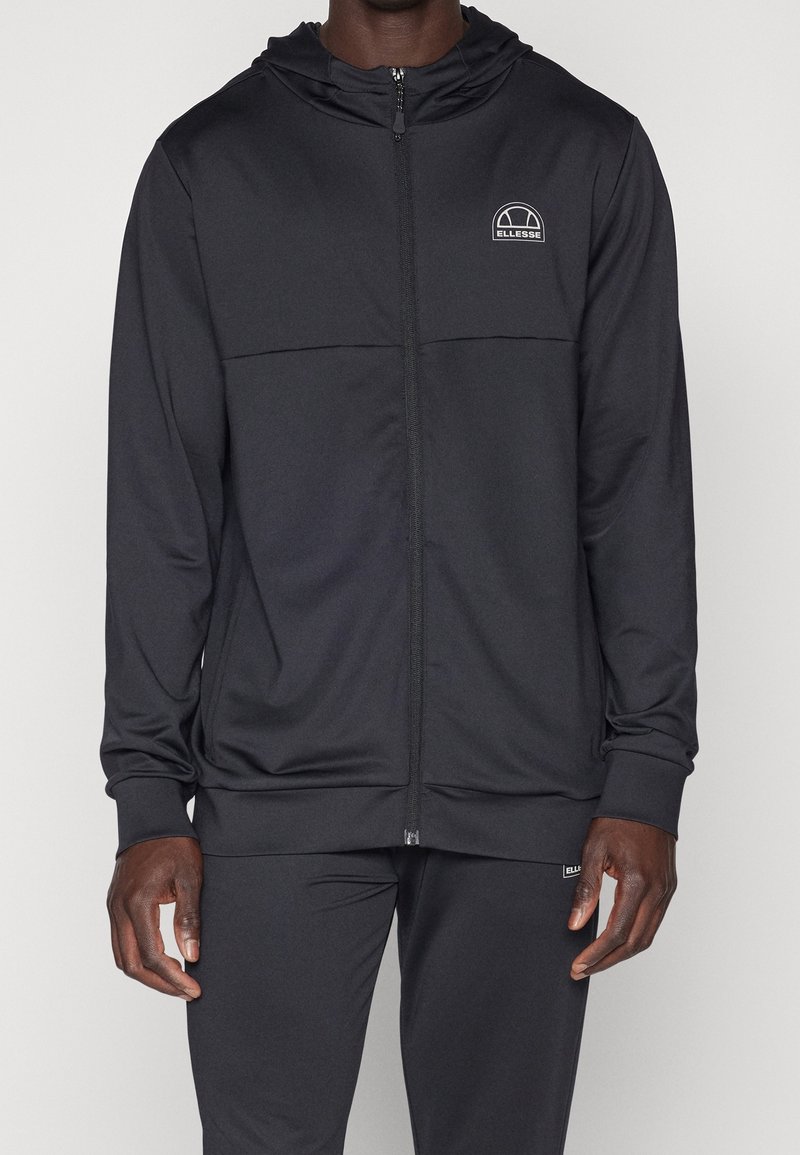 Haut à capuche zippé noir et pantalon assorti porté par une personne, avec le logo Ellesse sur la poitrine et la cuisse.