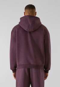 Haut à capuche violet avec une capuche spacieuse, des poignets et un ourlet côtelés, présentant une texture lisse et un design épuré. Associé à un bas de survêtement assorti.