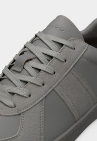 Gros plan sur une basket en cuir gris et daim avec des lacets gris assortis, une couture détaillée et le nom de la marque embossé sur la languette.