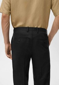 Pantaloni neri con una vestibilità su misura, dotati di due tasche posteriori con chiusura a bottone e una finitura liscia. Indossati con una camicia marrone.